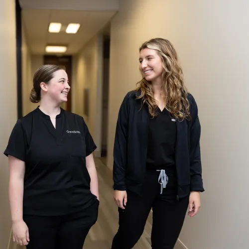 Two healthcare professionals walking down a modern hallway in scrubs, smiling at each other.
