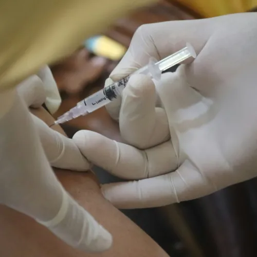 Gloved hands administer a vaccine injection into a person's arm using a syringe with a needle.