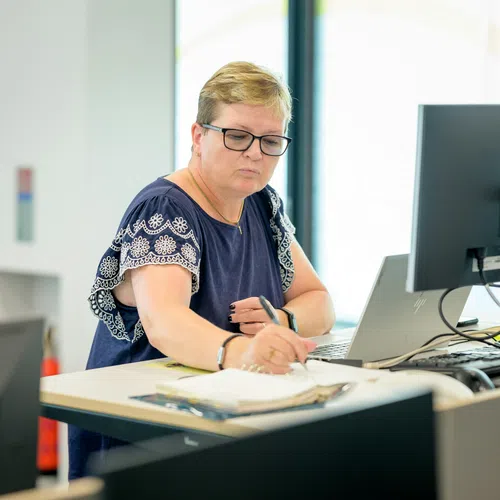 Woman in glasses working at a desk with computer writing in a notebook in a bright modern office.