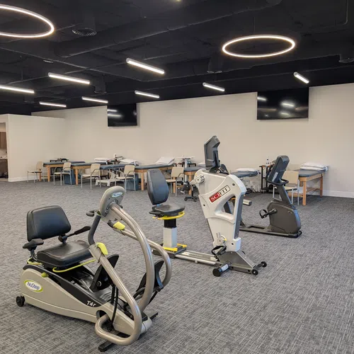 Physical therapy room with exercise bikes, treatment tables, and modern lighting fixtures on ceiling.