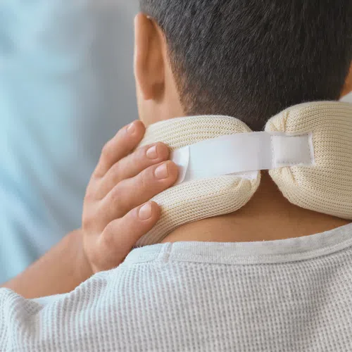 Person wearing a neck brace viewed from behind, holding the brace for support indoors in soft light.