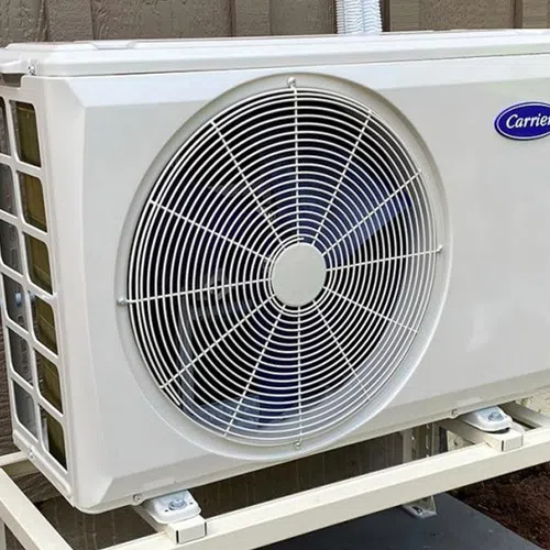 White Carrier outdoor air conditioning unit mounted on metal stand beside brown house wall.