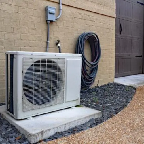Outdoor air conditioning unit beside a house with gravel pathway and utility hose.