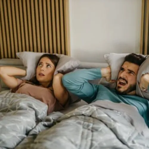 Frustrated couple in bed covering ears, struggling with noise disruption at night.
