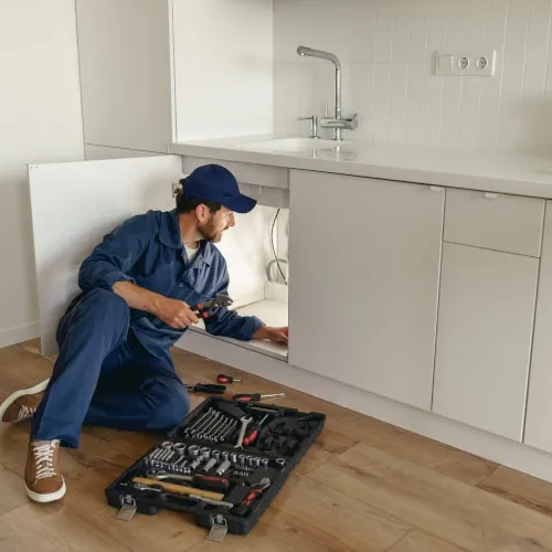 Plumber in blue overalls fixing kitchen cabinet pipes with tools on wooden floor under white countertop.