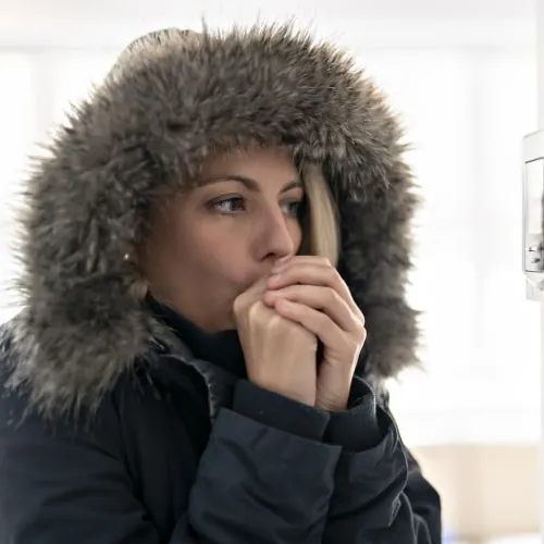 Woman in a winter coat with a fur hood warming her hands indoors near a thermostat.