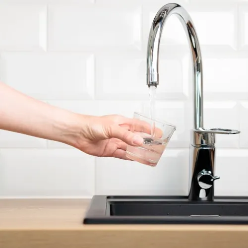 Hand holding a glass under a modern kitchen faucet filling it with water over a black sink and white tiled backsplash.