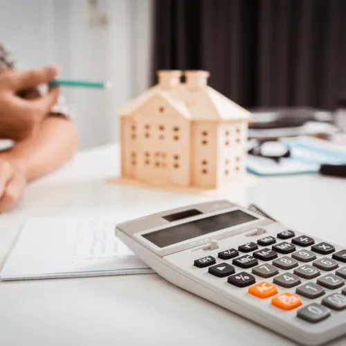 Person calculating home expenses with a calculator, a wooden house model, and paperwork on a white desk.