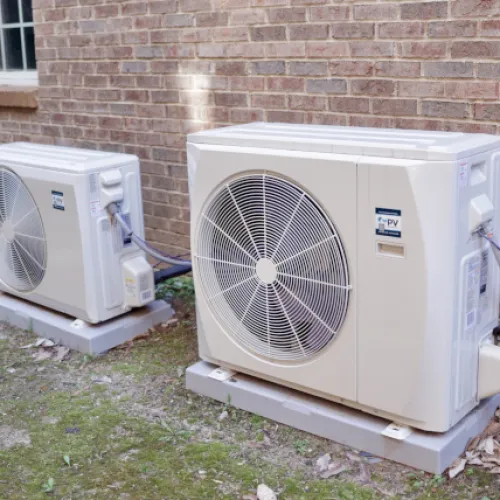 Two outdoor mini-split air conditioning units installed on concrete slabs beside a brick house wall with greenery around.