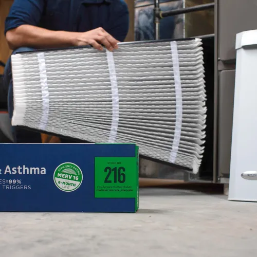 Person holding a pleated air filter near a furnace and an Allergy & Asthma air filter box labeled MERV 16 for 6-month use