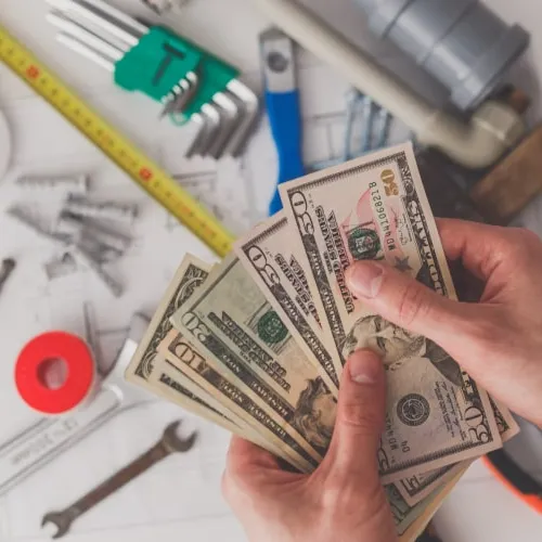 Person holding US dollar bills over plumbing tools and blueprints on a white table surface.