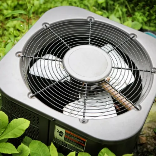 Outdoor air conditioning unit surrounded by green plants, showing the metal fan and protective grille.