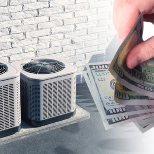 Hands holding US dollar bills near two outdoor air conditioning units mounted on a brick wall.