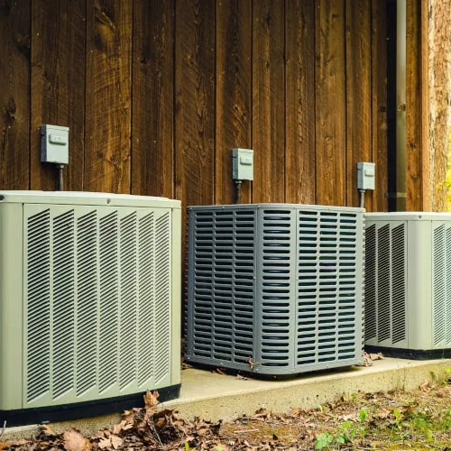 Three outdoor air conditioning units installed on concrete pad beside wooden wall in natural setting