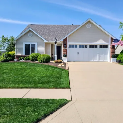 Single-story suburban house with a well-maintained lawn, two-car garage, and American flag on a sunny day