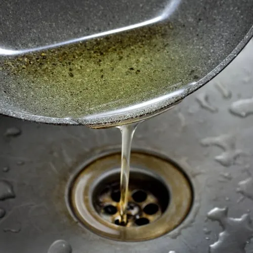 Used cooking oil being poured from a pan into a kitchen sink drain with water droplets around.