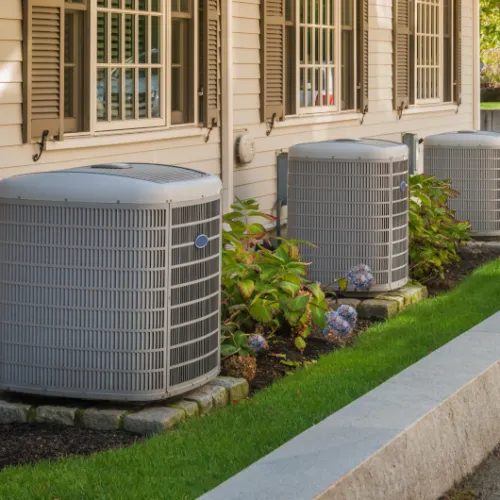 Three outdoor air conditioning units installed beside a beige house with green landscaping and stone bases.