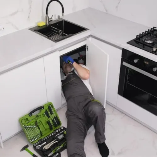 Plumber in uniform repairing kitchen sink pipes under the countertop with tools spread on the floor