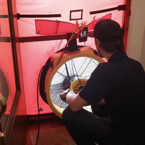 Man setting up large blower fan inside inflatable red air quality testing tent at home doorway.