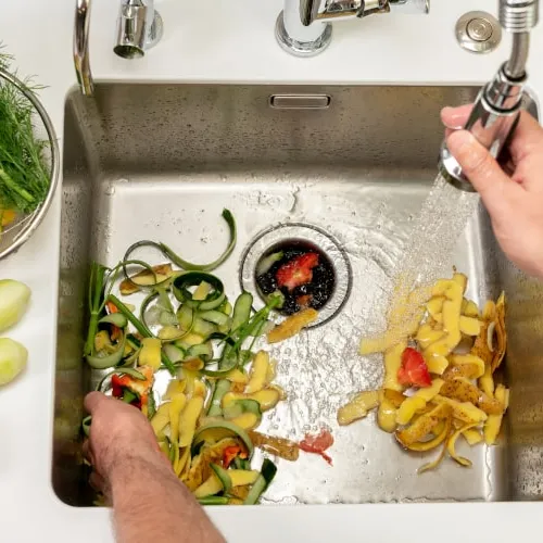 Person rinsing assorted vegetable peels and scraps in a stainless steel kitchen sink with running water.