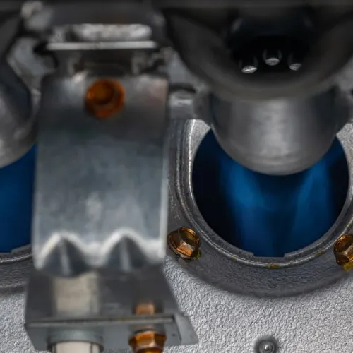 Close-up of blue flames burning inside a metal gas burner with bolts and mechanical parts visible.