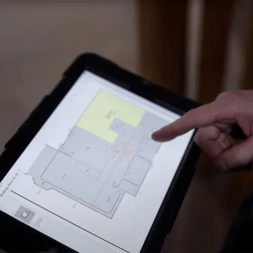 Person using a tablet to view and point at a digital floor plan layout in an indoor setting.