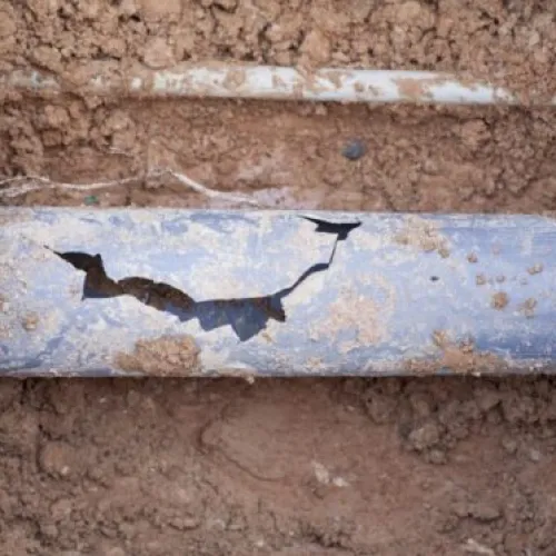 Damaged underground metal pipe with large cracks exposed in muddy soil during excavation work.