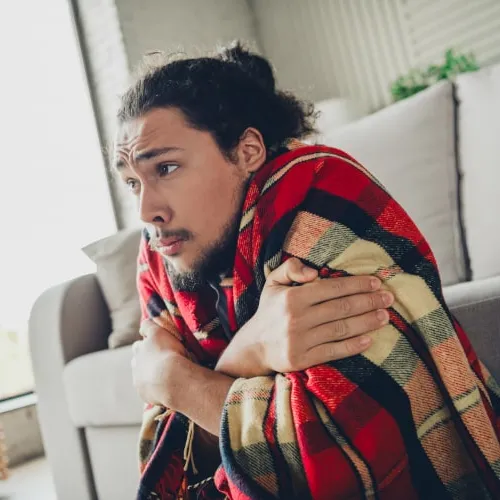 Man wrapped in a red plaid blanket sitting on the floor, looking cold and shivering indoors by the couch.