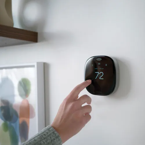 Person adjusting a modern black smart thermostat displaying 72 degrees mounted on a white wall.