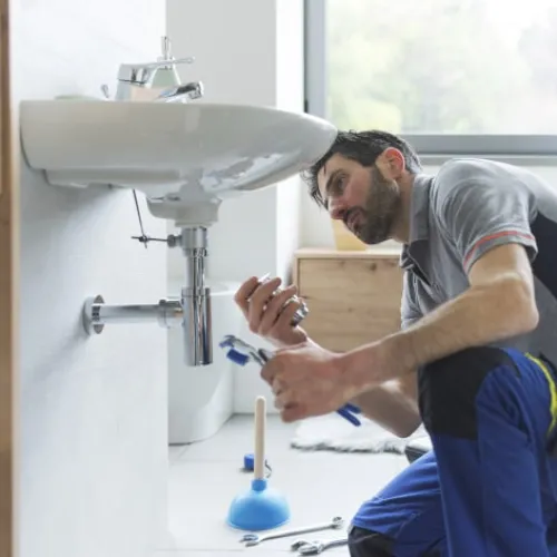 Plumber fixing a bathroom sink pipe using wrenches, with tools and plunger nearby in a modern bathroom.