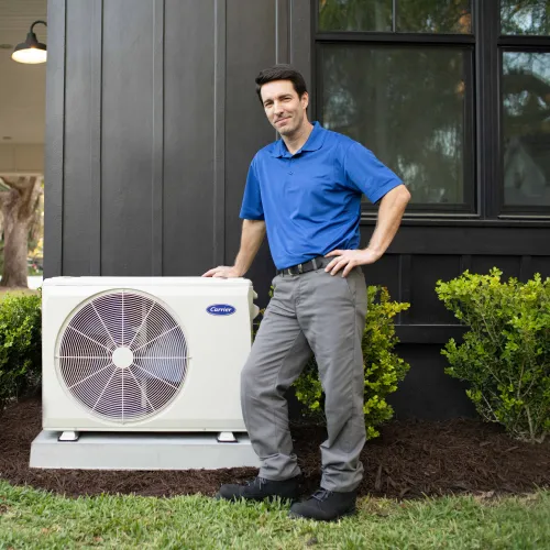 Technician standing next to a Carrier air conditioning unit outside a modern home with greenery.