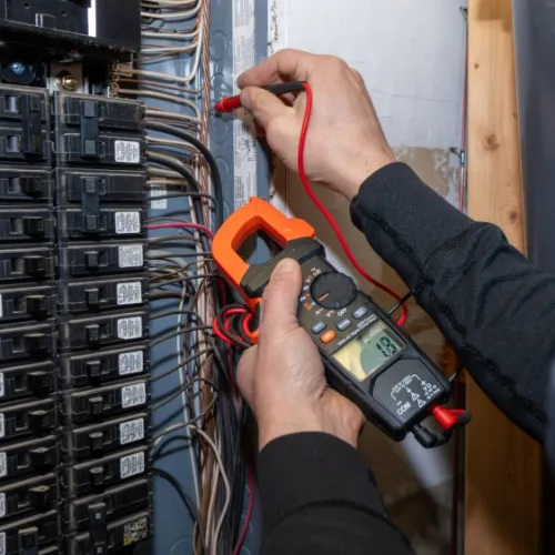 Electrician using clamp meter to test voltage in residential circuit breaker panel with multiple wires.