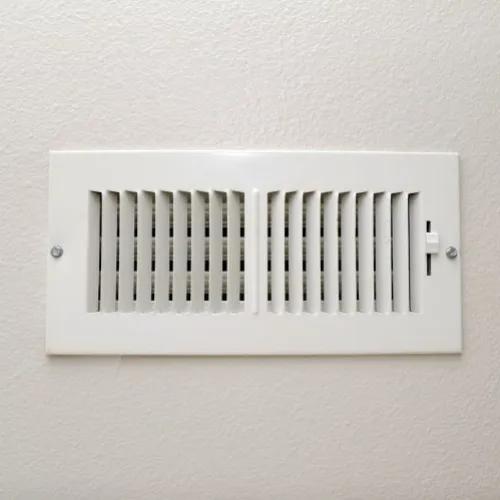 White rectangular air vent with adjustable louvers mounted on a beige wall with visible screws.
