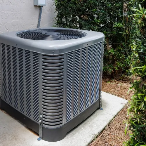 Outdoor central air conditioning unit installed on concrete slab beside house wall and green bushes.