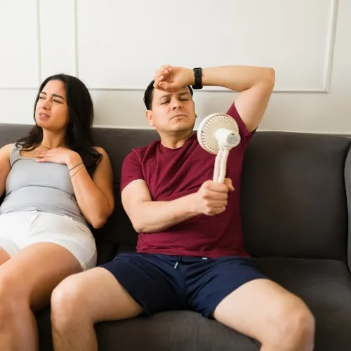 Man and woman sitting on couch using handheld fans to cool down feeling hot and uncomfortable indoors.