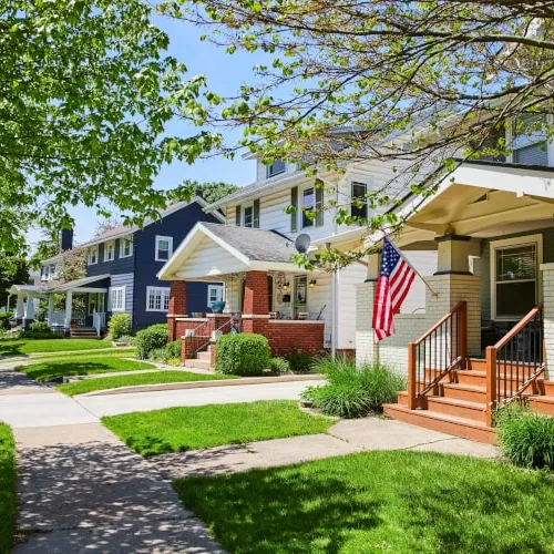 Row of suburban houses with front porches, green lawns, and an American flag on a sunny day.