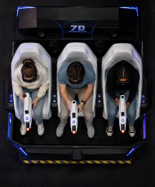 Top view of three people in a 7D interactive VR gaming pod holding white light guns and wearing VR headsets.