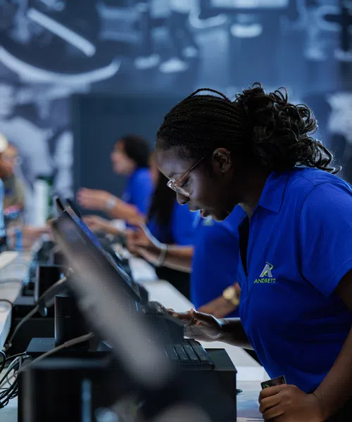 Woman in blue shirt helping customers at a busy tech service counter with multiple screens and devices.