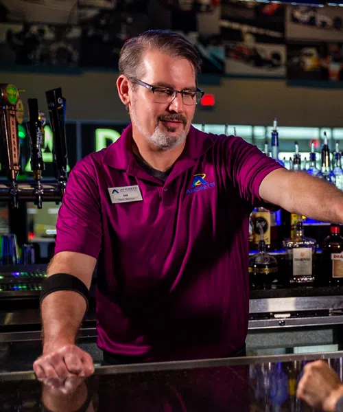 Bartender pouring a colorful cocktail at a bar counter for a female customer in a lively setting