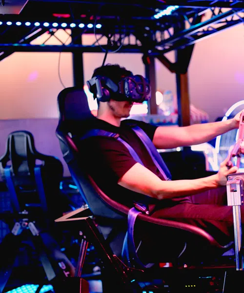 Man wearing VR headset plays interactive racing simulator game in a neon-lit arcade setup.