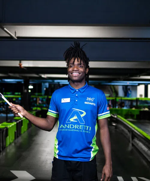 Young man in Andretti karting uniform smiling and holding a checkered flag in an indoor karting track.