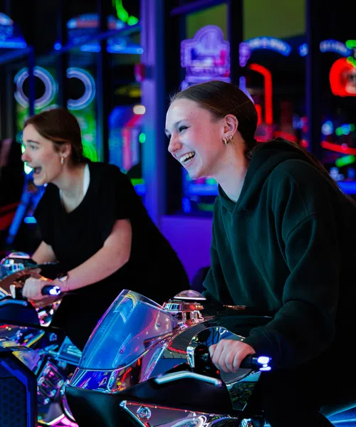 Two young women joyfully playing motorcycle arcade games surrounded by colorful neon lights.