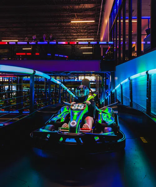 Person racing a green go-kart on an indoor track with neon blue and purple lights.