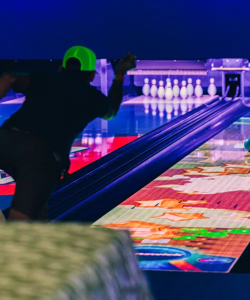 Person wearing neon green cap bowling on a colorful, illuminated lane with pins set up at the end under blue lighting.