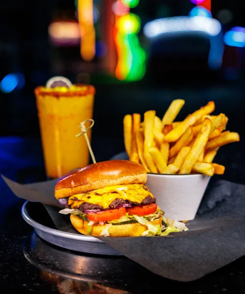 Cheeseburger with lettuce and tomato, cup of French fries, and a drink with colorful lights in background