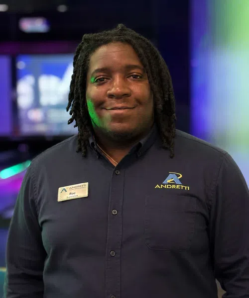 Smiling man with dreadlocks wearing a dark Andretti shirt and name tag Roc at an indoor event venue.