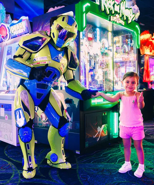 Child giving thumbs up while holding hand with yellow and blue robot in vibrant arcade game room.