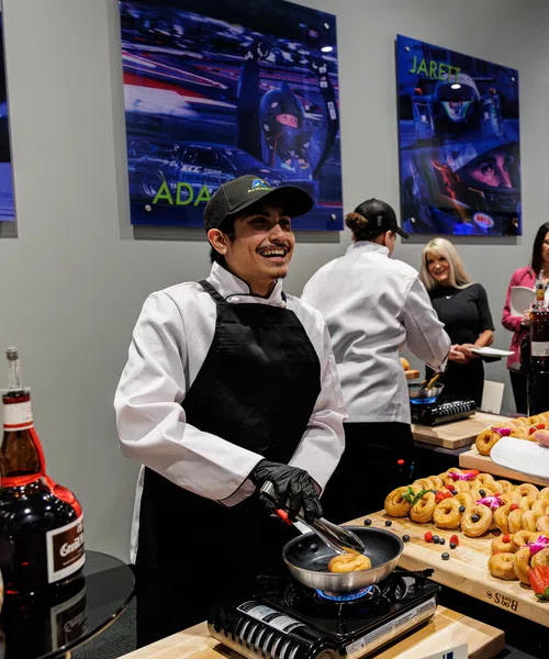 Chef wearing black apron serving pastries to guests at an indoor event with vibrant wall art and refreshments.