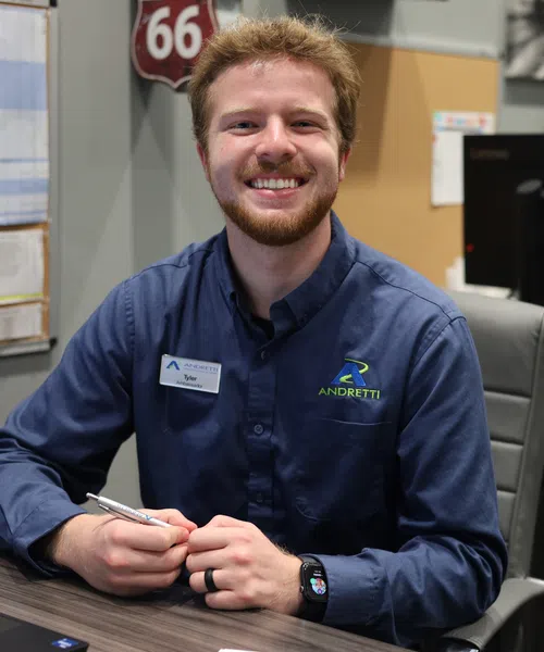 Smiling man in a blue Andretti uniform sits at an office desk holding a pen, surrounded by computers and office items