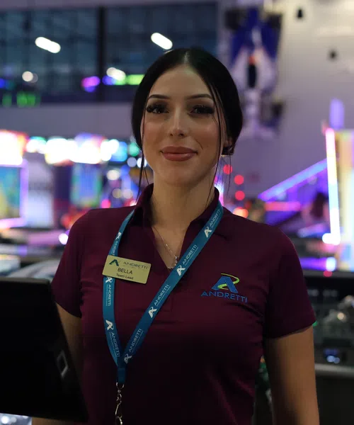 Smiling female bartender in purple Andretti shirt stands behind bar with colorful lights in background.